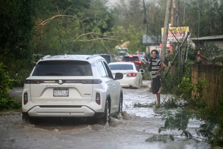 A white car behind many other cars drives through flood waters as a Black man watches
