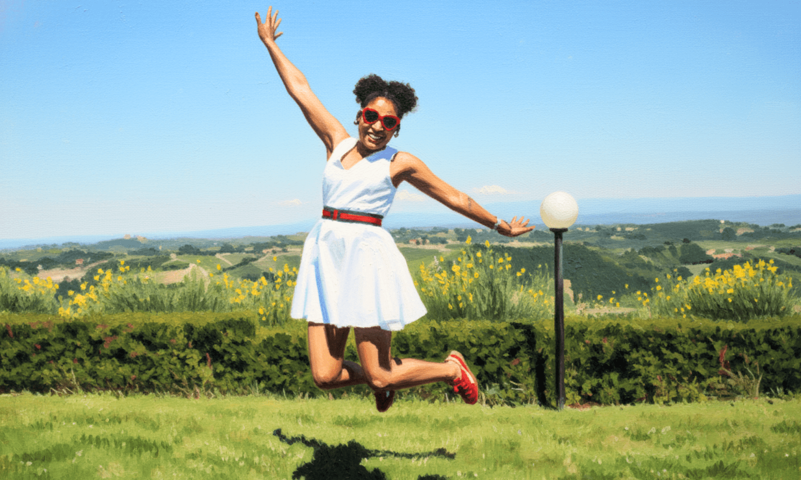 A young Black woman wearing a white dress and red-framed heart-shaped sunglasses jumps up in a large field