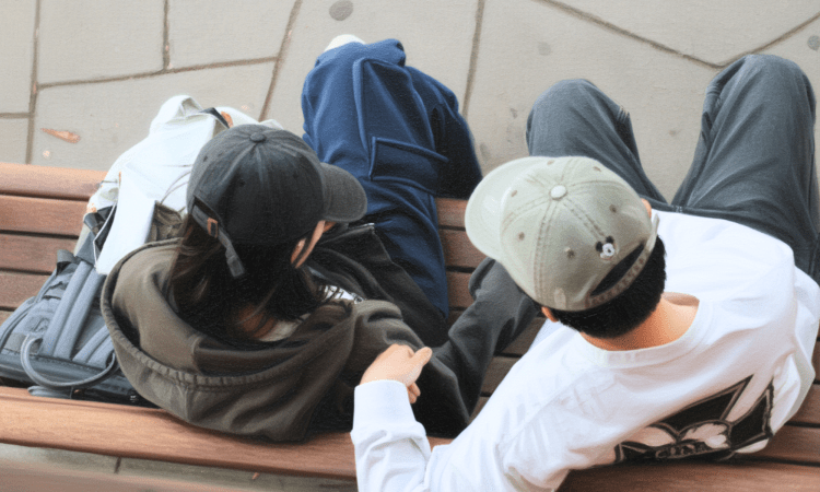 Aerial view of two young people sitting on a bench outdoors. Both wear caps and they have a backpack beside them