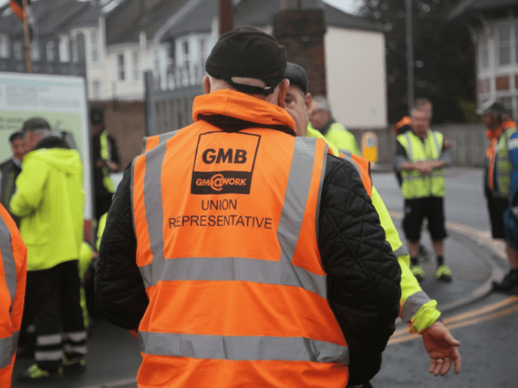 An older white man with his back to the camera wears a hi-vis vest that reads "GMB Union Representative." He speaks to another older while man with other workers in the background