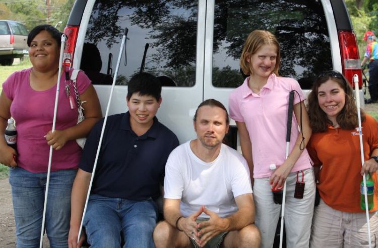 A group of people smile in front of a van, with four of them holding white canes