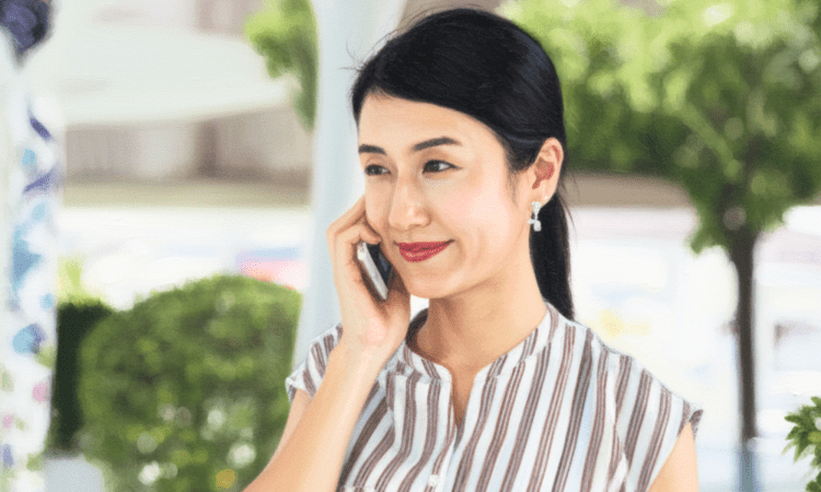 Asian woman wearing a pony tail is similing as she talks on the phone outdoors