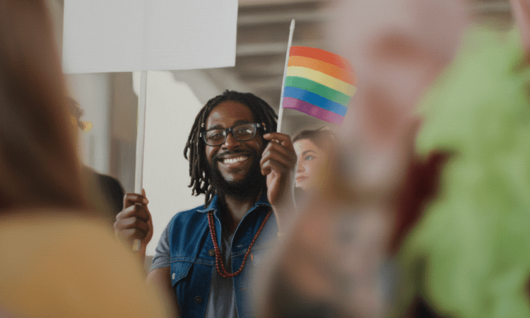 A Black man with dreads is smiling broadly, holding a pride flag