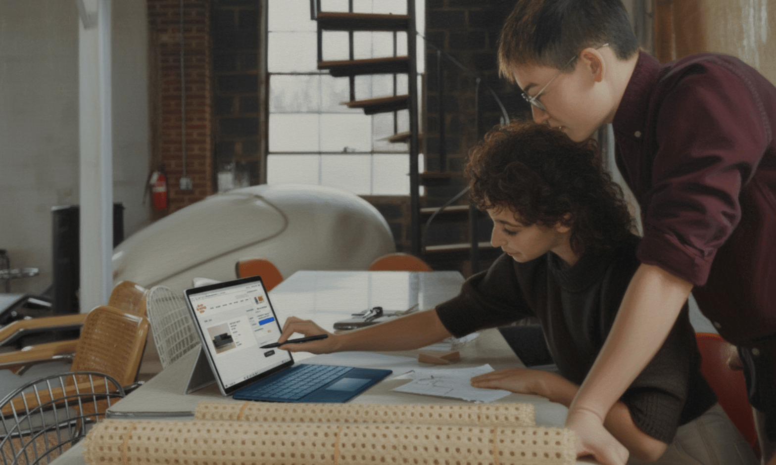A woman uses a stylus to pint to an image on a laptop while a man stands over her and watches
