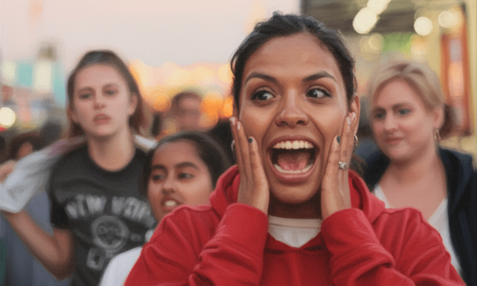 A young woman of colour holds her hands to her cheeks, her mouth open in happy surprise. Three young women stand behind her