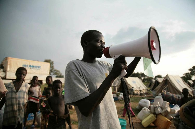 A Black man smiles while talking into a megaphone, with children and UNHCR tents in the background