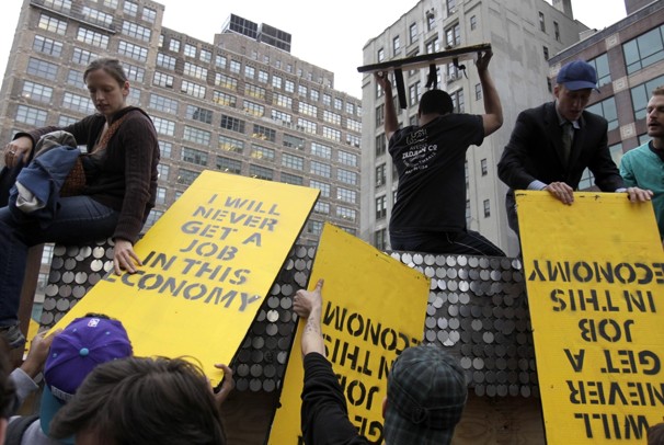 Young people sit high above a crowd, grabbing yellow signts that say: I will never get a job in this economy