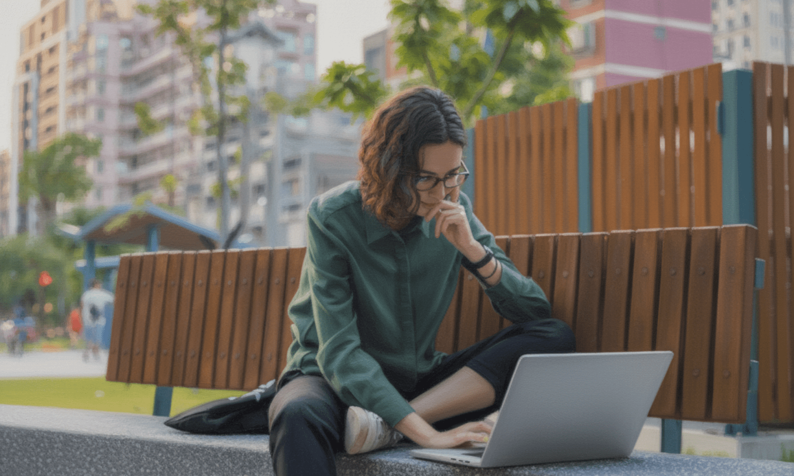 A white woman sits crossed leg on a park bench smiling, working on her laptop