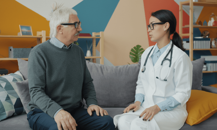 A white elderly man with white hair sits on a couch beside an Asian woman doctor, who is speaking