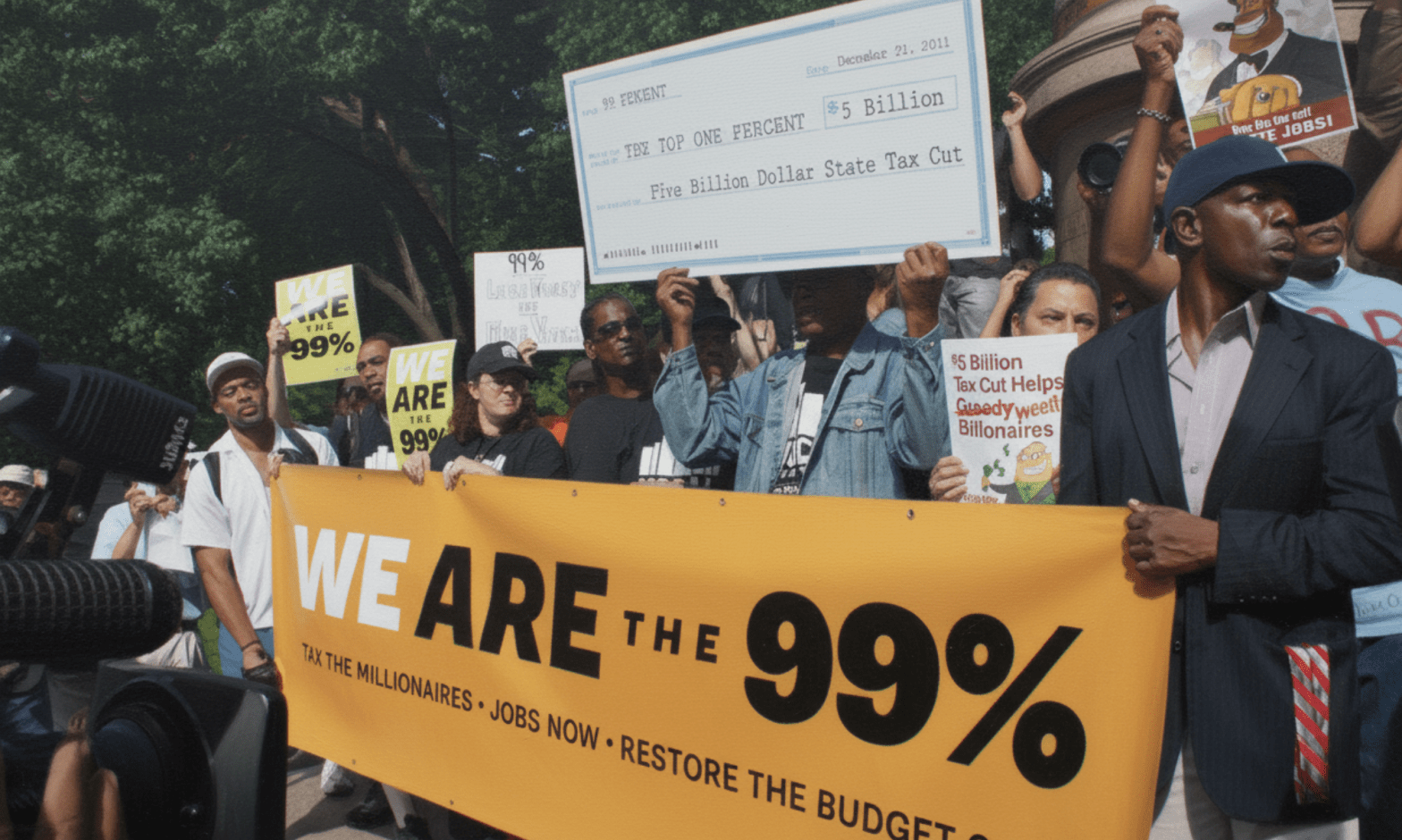 Black and white people hold a large yellow sign saying: WE ARE THE 99%. One person holds up a giant cheque, which says: Five Billion Dollar State Cut
