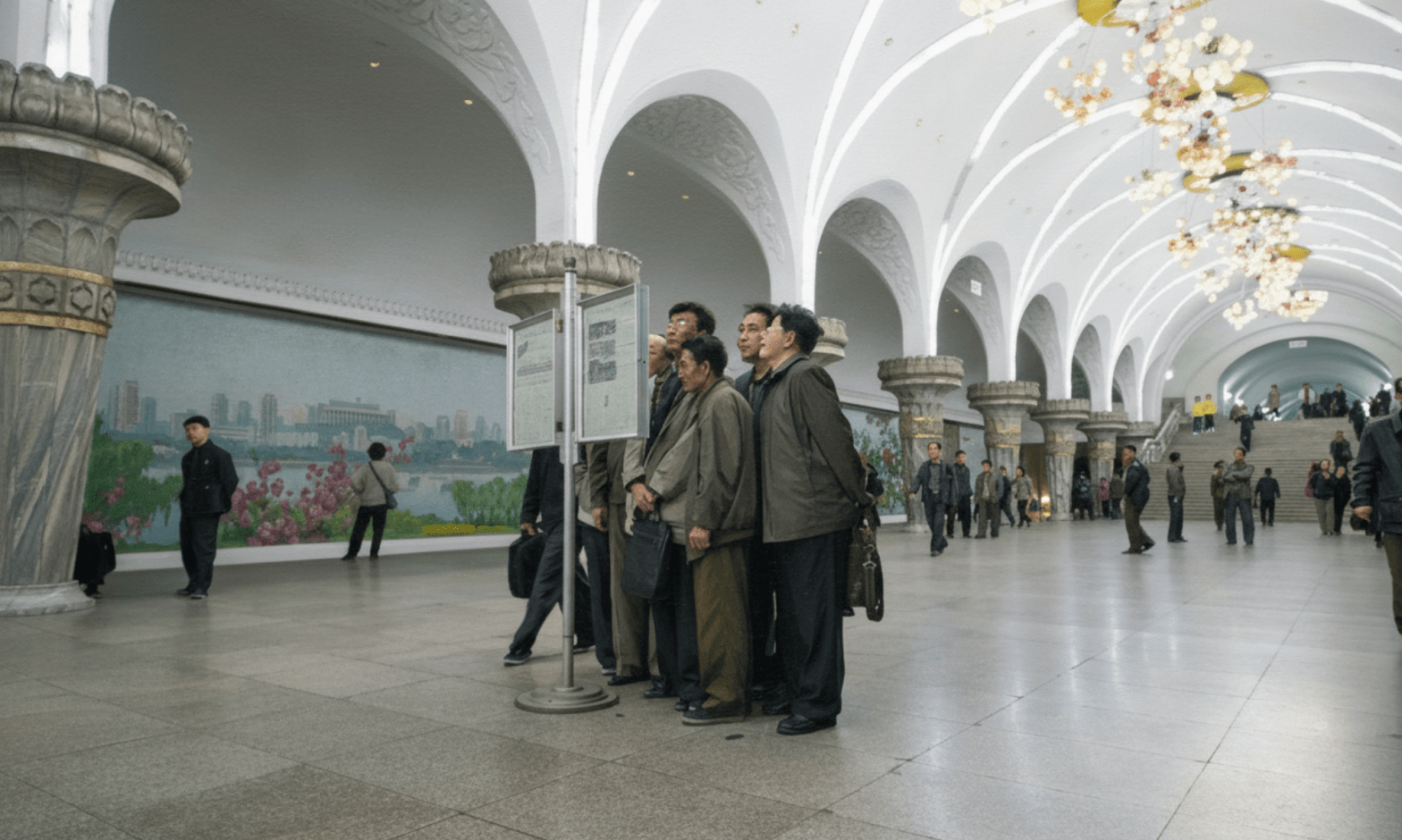 In the Metro of North Korea, a group of men read a bulletin board, with people walking in the background