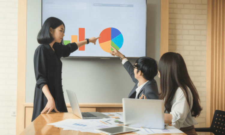 An Asian woman stands, pointing to a presentation of graphs, while two other people look up